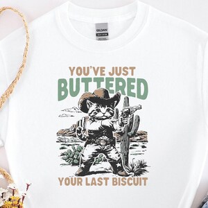 May include: White t-shirt with a graphic of a cat dressed as a cowboy, holding a cactus and a gun. The text reads "YOU'VE JUST BUTTERED YOUR LAST BISCUIT" in green and brown. The shirt is paired with denim shorts.