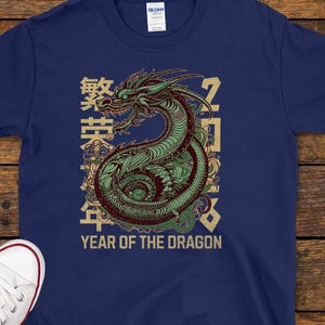 Puede incluir: Camiseta azul marino con un diseño de dragón verde y rojo y el texto "YEAR OF THE DRAGON". El diseño incluye caracteres chinos. Zapatillas de lona blancas con ribete rojo están al lado de la camiseta.