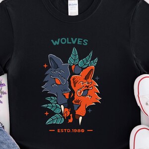 Pode incluir: T-shirt preto com um gráfico de duas cabeças de lobo, uma azul e outra laranja, com folhas verdes e o texto "WOLVES ESTD.1980". A camisa é combinada com calções de ganga azuis e ténis brancos.