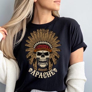 May include: Black t-shirt featuring a skull wearing a Native American headdress. The design includes the words "NATIVE AUTHENTIC WARRIORS APACHE" in a vintage style. The shirt is made of a soft material.