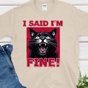 Op de afbeelding: Beige t-shirt met een afbeelding van een zwarte kat met open mond, tegen een rode achtergrond. De tekst "I SAID I'M FINE!" staat in rode letters boven en onder de kattenafbeelding. Een blauwe spijkerbroek ligt naast het t-shirt.