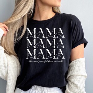 以下が含まれることがあります： 白文字で「MAMA」と3回繰り返された黒いTシャツ。下に「The most powerful force on earth」というフレーズがあります。 シャツはリブ編みで、半袖です。