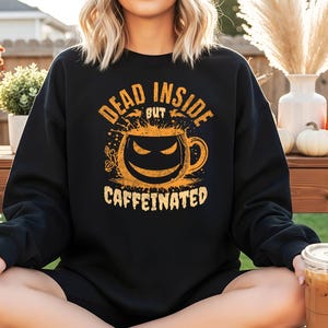 Könnte beinhalten: Schwarzes Sweatshirt mit dem Text "DEAD INSIDE BUT CAFFEINATED" in Orange. Die Grafik zeigt ein Kürbisgesicht in einer Kaffeetasse. Die Person trägt braune Hausschuhe und hält ein Getränk mit einem Strohhalm.