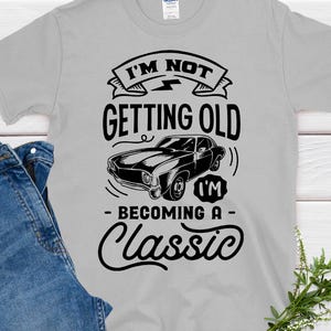 Może przedstawiać: Jasnoszara koszulka z napisem "I'M NOT GETTING OLD I'M BECOMING A CLASSIC" i grafiką klasycznego samochodu. Koszulka jest połączona z niebieskimi dżinsami. Gałązka zieleni znajduje się w prawym dolnym rogu.