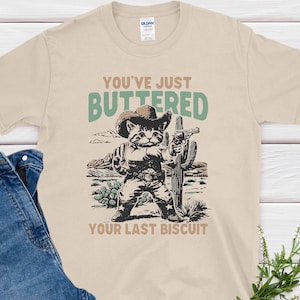 May include: Beige t-shirt with a graphic of a cat in a cowboy hat holding a gun and cactus. Text on the shirt reads "You've Just Buttered Your Last Biscuit." Blue denim jeans are folded next to the shirt.
