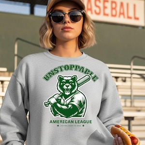 Könnte beinhalten: Ein hellgraues Sweatshirt mit einem grünen Aufdruck eines Bären, der einen Baseballschläger hält. Der Text "UNSTOPPABLE" steht über dem Bären, darunter "AMERICAN LEAGUE". Das Sweatshirt hat auch den Text "ESTABLISHED IN 2023".