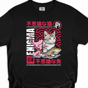 Puede incluir: Camiseta negra con un diseño de inspiración japonesa. El diseño incluye un gato, un pez, flores de cerezo y texto japonés. La palabra "ENIGMA" está impresa verticalmente en el lado izquierdo.