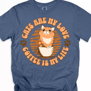 Op de afbeelding: Blauw t-shirt met een cirkelvormige afbeelding met een cartoonkat in een koffiekopje. De tekst "CATS ARE MY LOVE COFFEE IS MY LIFE" staat in oranje rond de kat. Aan weerszijden van de afbeelding staan pootafdrukken.