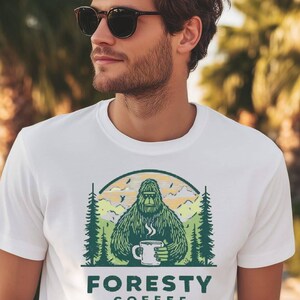 以下が含まれることがあります： 白いTシャツ。ビッグフットがコーヒーカップを持っているグラフィックで、緑色の「FORESTY COFFEE」の文字が入っています。デザインには木、山、虹が含まれています。Tシャツが着用されています。