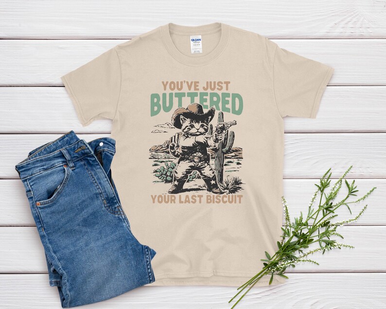 May include: Beige t-shirt with a graphic of a cat in a cowboy hat holding a gun and cactus. Text on the shirt reads "You've Just Buttered Your Last Biscuit." Blue denim jeans are folded next to the shirt.