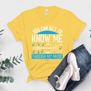 K&ouml;nnte beinhalten: Gelbes T-Shirt mit dem Text "YOU CAN GET TO KNOW ME THROUGH MY MUSIC" in Wei&szlig; und Blau. Das Design umfasst Noten und eine Gitarre. Das Shirt wird mit einer blauen Jeans und wei&szlig;en Turnschuhen kombiniert.