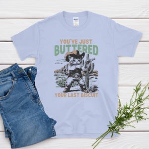 May include: Light blue t-shirt with a graphic of a cat in a cowboy hat holding a cactus, with the text "You've Just Buttered Your Last Biscuit." The shirt is paired with blue jeans.