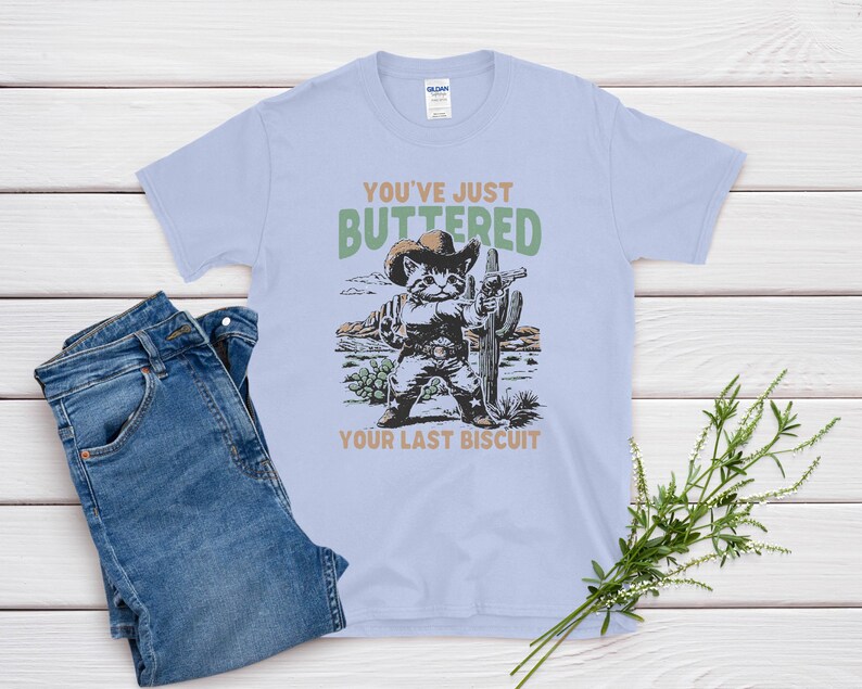 May include: Light blue t-shirt with a graphic of a cat in a cowboy hat holding a cactus, with the text "You've Just Buttered Your Last Biscuit." The shirt is paired with blue jeans.