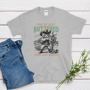 May include: A light gray t-shirt with a graphic of a cat dressed as a cowboy, holding a gun and standing in front of a cactus. The text on the shirt reads "YOU'VE JUST BUTTERED YOUR LAST BISCUIT."