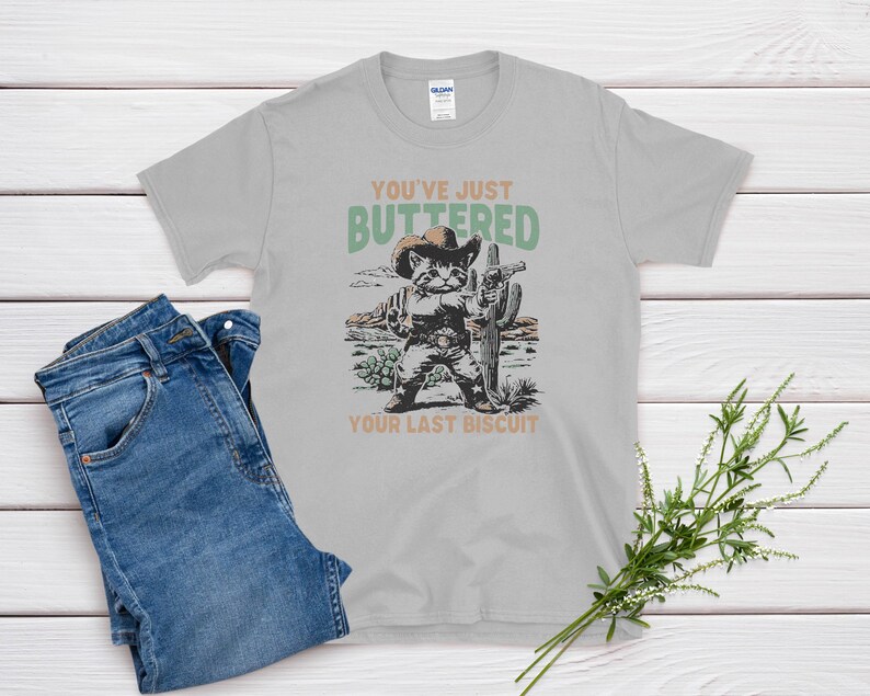 May include: A light gray t-shirt with a graphic of a cat dressed as a cowboy, holding a gun and standing in front of a cactus. The text on the shirt reads "YOU'VE JUST BUTTERED YOUR LAST BISCUIT."
