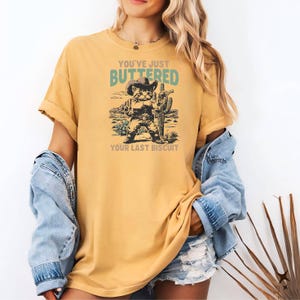 May include: Mustard yellow t-shirt featuring a graphic of a cat dressed as a cowboy, holding a gun, and standing in front of a cactus. The text on the shirt reads "YOU'VE JUST BUTTERED YOUR LAST BISCUIT."