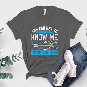 K&ouml;nnte beinhalten: Ein graues T-Shirt mit dem Text "YOU CAN GET TO KNOW ME THROUGH MY MUSIC" in Wei&szlig; und Blau. Das Design umfasst Noten und eine Gitarren-Grafik. Das Shirt wird mit blauen Jeans und wei&szlig;en Turnschuhen kombiniert.