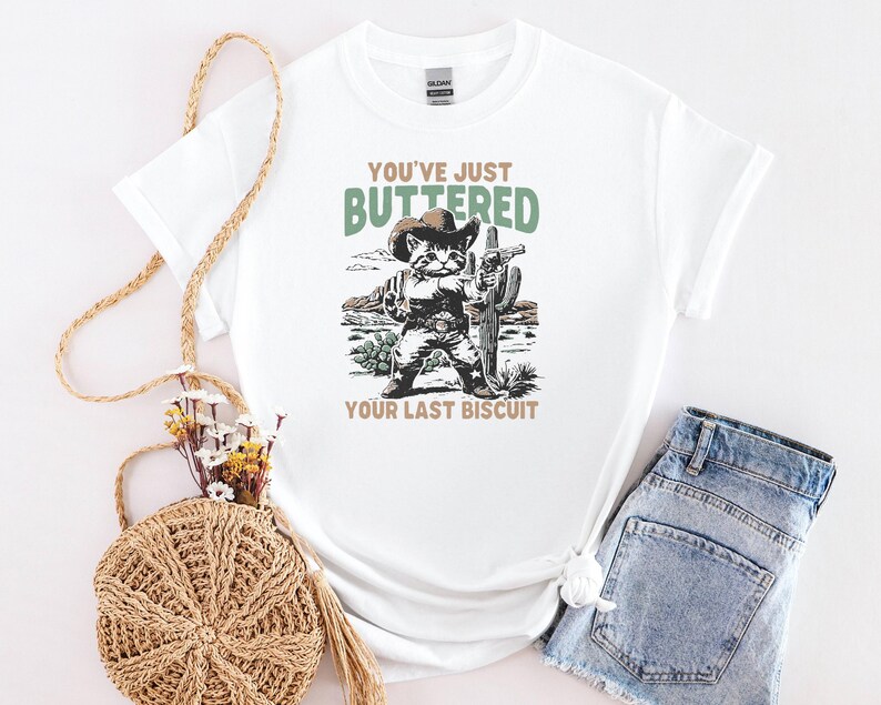 May include: White t-shirt with a graphic of a cat dressed as a cowboy, holding a cactus and a gun. The text reads "YOU'VE JUST BUTTERED YOUR LAST BISCUIT" in green and brown. The shirt is paired with denim shorts.