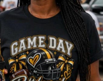 May include: A black short-sleeved t-shirt featuring a graphic print with the text GAME DAY Pittsburgh kind of girl. The design includes a black and yellow football helmet, a brown football, and yellow and black cheerleading bows with hearts.