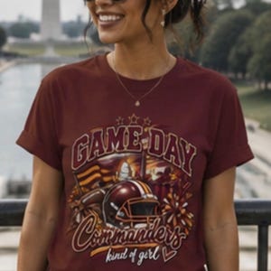 May include: A maroon short-sleeve t-shirt featuring a graphic print with the text "GAME DAY" and "Commanders kind of girl" alongside a football helmet, a football, and a flag. The design includes stars and a small heart icon below the text.