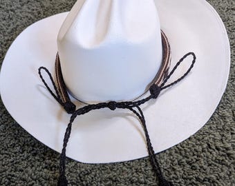 May include: A white cowboy hat with a brown and black hatband and a black chin strap with white tassels. The hat has a wide brim and a rounded crown. The hat is made of a stiff material.
