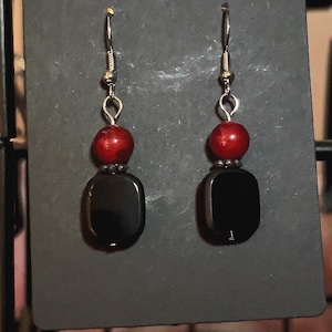 May include: A pair of dangle earrings featuring a red bead above a large, black, rectangular bead. The earrings have silver-colored ear wires and are displayed on a black card.