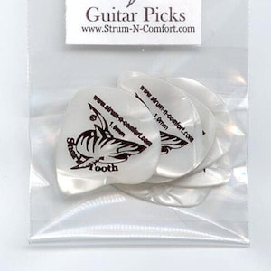 May include: Guitar picks in a clear plastic package. The picks are white with a shark tooth logo and the text "Shark Tooth". The packaging also displays the text "Guitar Picks" and a website address.