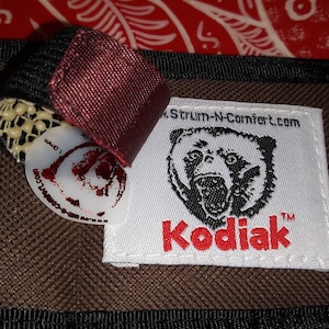 May include: Close-up of a brown and black guitar case with a label that reads "Kodiak" and features a bear illustration. The case has a black trim and a burgundy fabric tag. A white guitar pick with the text "www.strum-n-comfort.com" is visible.