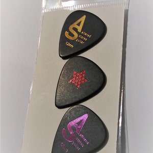 May include: Three black guitar picks in a clear plastic package. Each pick has a different design: one with a gold "SA" logo and "altered states guitar" text, one with a red star, and one with a pink "SA" logo.