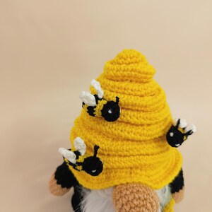 May include: A handmade crochet gnome with a yellow beehive-shaped hat and a white beard. The hat is adorned with three small, black and yellow bees. The gnome's body is black and white, with tan hands.