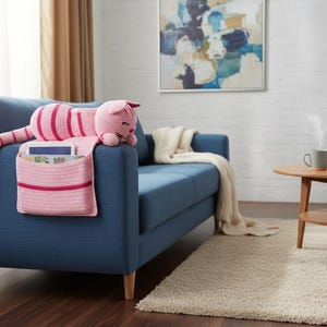 May include: A pink and red crocheted cat-shaped couch organizer with a pocket for storage. The cat is lying on a blue couch. The organizer has a striped body and a smiling face. A tablet and books are in the pocket.