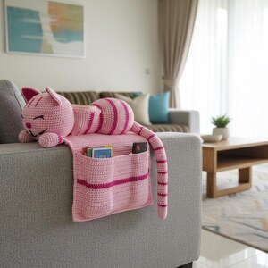 May include: A pink crocheted cat-shaped couch organizer with pockets. The cat has a smiling face, ears, and a striped tail. The organizer is draped over the arm of a gray couch, with pockets holding cards and a remote.