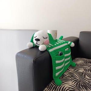 Könnte beinhalten: Gehäkelter Hunde-Armlehnen-Organizer in Grün und Weiß gestreift. Der Hund hat einen weißen Kopf, rosa Wangen und geschlossene Augen. Er hat Taschen zur Aufbewahrung und hält einen weißen Becher.