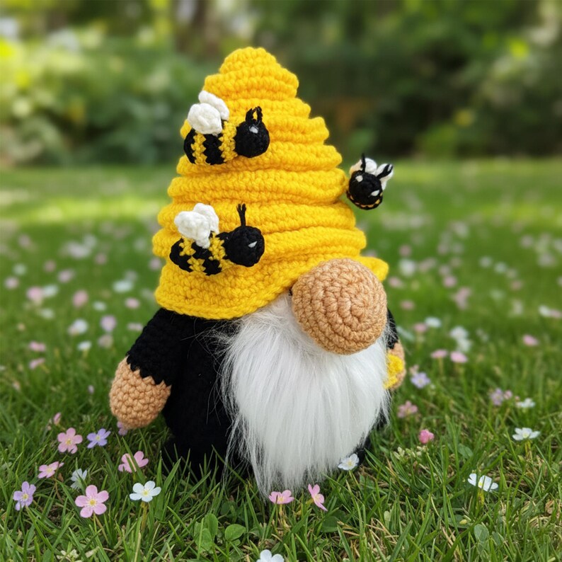 May include: A handmade crochet gnome with a yellow beehive hat and a white beard. The gnome has a black body and tan arms. Three small bees are attached to the hat. The gnome is sitting in a grassy area with small flowers.