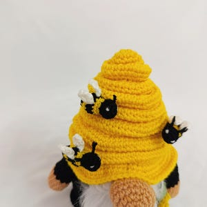May include: A handmade crochet gnome with a yellow beehive hat and three attached bees. The gnome has a white beard, black and yellow body, and tan arms and feet. The gnome is a decorative item.