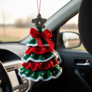Könnte beinhalten: Handgefertigter Weihnachtsbaum-Autoschmuck. Der Baum ist aus grüner und roter Wolle gehäkelt, mit weißen Perlen verziert und mit einem silbernen Stern gekrönt. Eine rote Schleife ist unter dem Stern gebunden.