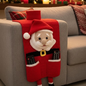 May include: A red crocheted Santa Claus couch organizer with pockets for remote controls. The Santa face is white and tan with a red hat and black belt. The organizer is draped over the arm of a gray couch, with Christmas decorations in the background.