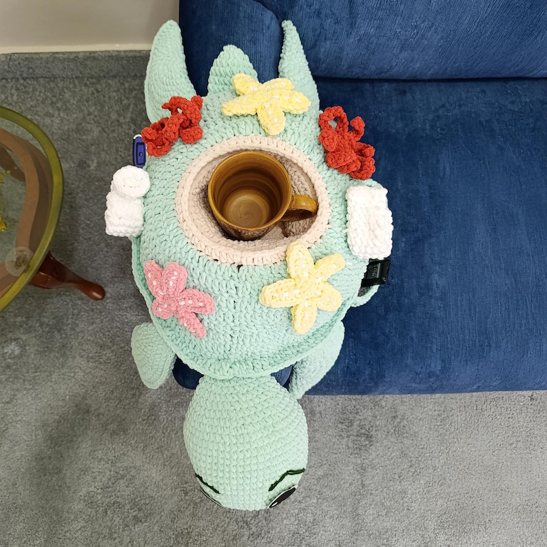 May include: A handmade, light green crocheted turtle-shaped table tray with a built-in cup holder. The tray is decorated with crocheted starfish in red, pink, and yellow. A brown mug sits in the center. The tray is designed to sit on the arm of a sofa.