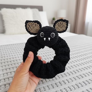 May include: A black crochet hair scrunchie with a bat design. The bat has brown ears, white fangs, and round eyes. The scrunchie is held in a hand and is set against a soft background.