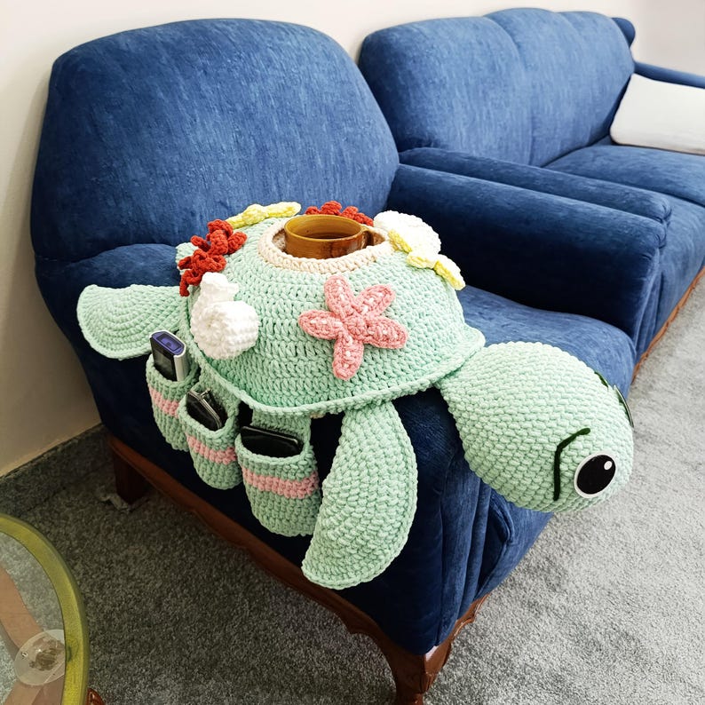 May include: A crocheted turtle-shaped sofa armrest organizer in shades of green, with a cup holder and pockets for remote controls and phones. The turtle has a pink starfish and white and yellow floral accents.
