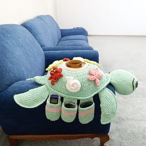 May include: A handmade, light green crocheted turtle-shaped sofa armrest organizer. It features a central cup holder, decorative sea-themed embellishments, and pockets for remote controls and other small items. The turtle's head and flippers extend from the main body.
