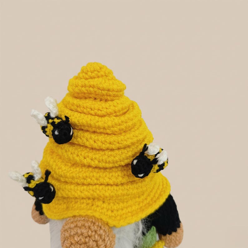 May include: A handmade gnome with a yellow crocheted beehive-shaped hat and three attached bee figures. The gnome has a white beard, a tan nose, and black and yellow striped bee bodies with white wings. The background is a solid beige color.
