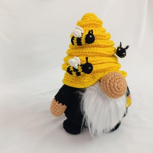 May include: A handmade crochet gnome with a yellow beehive hat and a white beard. The gnome has a black body, tan hands and nose, and is decorated with three black and yellow bees. The gnome is a decorative item.