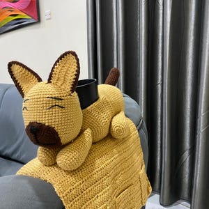 Puede incluir: Una almohada hecha a mano con forma de perro, tejida a ganchillo, en tonos beige y marrón. El perro está tumbado sobre una manta de ganchillo beige a juego con dos bolsillos. La almohada está colocada sobre un sofá gris, con un objeto cilíndrico negro en la espalda del perro.