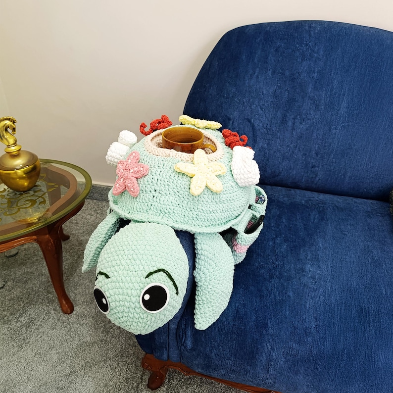 May include: A light green crocheted turtle-shaped armrest cover with a built-in cup holder. The turtle's shell is decorated with pink, white, and yellow crocheted starfish and coral. The turtle has large, expressive eyes and flippers.