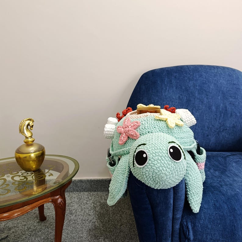 May include: A handmade, light teal crochet turtle-shaped bag with large black eyes and a floral crown. The turtle has a smiling face and is decorated with pink, yellow, and white crochet flowers. The bag is resting on a blue armchair.