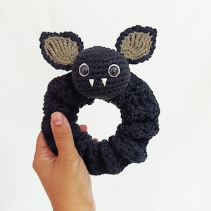 May include: A black crochet scrunchie with a bat design. The bat has large white eyes, small white fangs, and tan-colored ears. The scrunchie is held in a hand against a white background. The scrunchie is a Halloween-themed hair accessory.