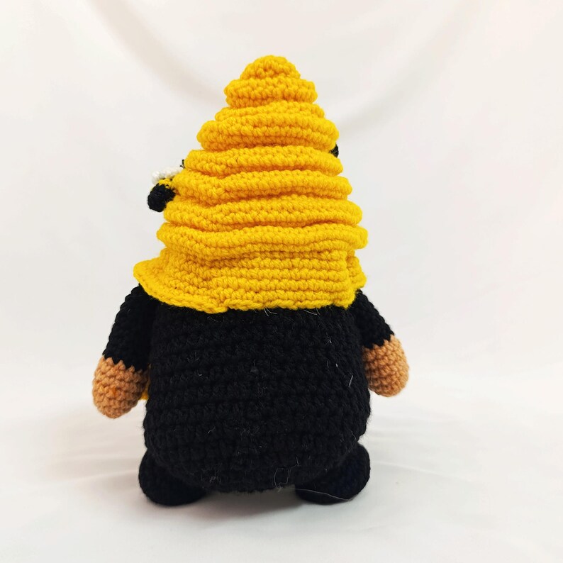 May include: A handmade crochet gnome with a yellow beehive hat and black body. The gnome has small tan hands and feet. The gnome is standing on a white surface.