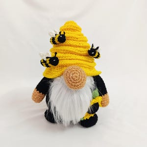 May include: A handmade crochet gnome with a yellow beehive hat and a long white beard. The gnome has black and yellow accents, with small bee embellishments. The gnome's nose is a light brown color. The overall design is whimsical and decorative.