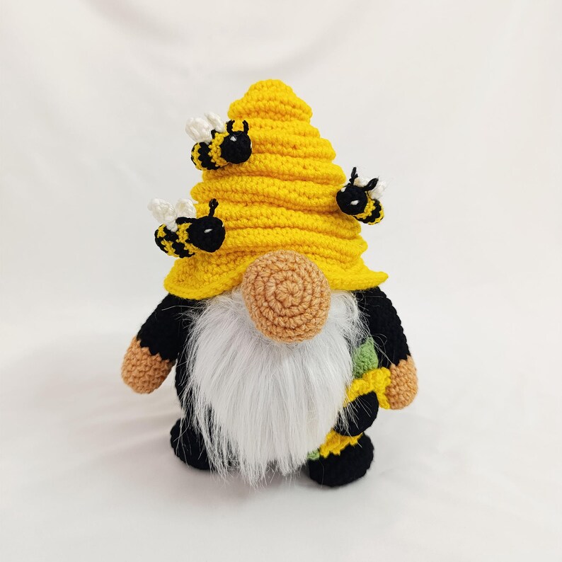 May include: A handmade crochet gnome with a yellow beehive hat and a long white beard. The gnome has black and yellow accents, with small bee embellishments. The gnome's nose is a light brown color. The overall design is whimsical and decorative.